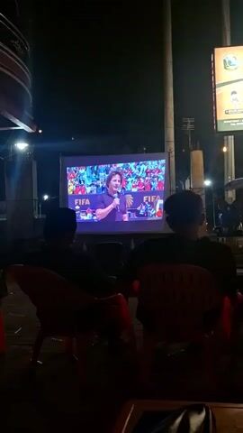 The video shows a group of people sitting in chairs, watching a large projection screen outdoors at night. On the screen, a man is speaking, with the FIFA logo visible. The man appears to be in a stadium with a crowd in the background. The people watching are seated and facing the screen, with their backs to the camera for the most part. The environment appears to be a casual outdoor setting, possibly a cafe or gathering place, with some structural elements and lighting visible. The overall atmosphere seems relaxed, with people gathered to watch the event on the screen.