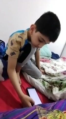 A young boy with dark hair and a blue and tan shirt kneels on a bed. He is looking down at a white screen on a phone he holds in his right hand. A person’s hand with a gold and patterned sleeve reaches into the frame and gently strokes the boy’s hair. The bed is covered with a patterned duvet in shades of white, green, pink, and blue, and a red sheet is visible in the foreground. The background is a plain white wall.