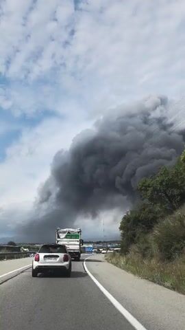 A white car with its taillights illuminated drives on a highway, following a large green and white truck. Thick, dark gray smoke billows into the sky on the right side of the road, obscuring the horizon. Overhead power lines stretch across the frame against a cloudy blue sky. Lush green trees and bushes line the right side of the road. Another white car is visible in the distance on the highway.