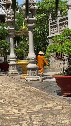 The video shows a monk walking through a courtyard. The courtyard is paved with light brown bricks. Two tall, gray stone pillars with ornate bases and lanterns on top stand on either side of a large golden vase. The monk, wearing an orange robe and a conical hat, walks behind the vase. There are potted plants with small trees on either side of the walkway, and a building with gray stone walls and ornate carvings is visible in the background. The environment suggests a Buddhist temple or monastery.