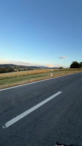 This is a first-person perspective video of a bicycle ride on a paved road in Ufhusen, Switzerland. The scene is set in a rural landscape with rolling hills, fields of dry grass, and distant mountains under a clear blue sky with a few wispy clouds. The time of day appears to be late afternoon or early evening, as indicated by the soft, warm light and the long shadows.

The road is asphalt with white lane markings. A single white marker post is visible on the left side of the road. In the distance, several buildings are scattered across the landscape, including a house with a dark roof and a barn or agricultural building. A white truck or van is parked on the right side of the road near one of the buildings. Trees are also scattered throughout the scene, some with full green foliage and others appearing more bare.

The camera is mounted on the handlebars of the bicycle, showing a portion of the bike's frame and controls in the foreground. The overall mood is peaceful and serene, evoking a sense of freedom and enjoyment of the natural scenery and outdoor activity. There is no visible text in the video.