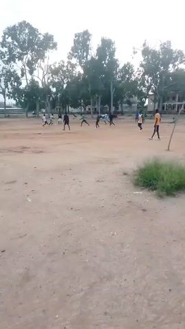 The video shows an outdoor sports field with several people playing a game, likely soccer. The field is predominantly dirt with some patches of grass. In the background, there are numerous trees and some buildings. The sky is overcast. The players are scattered across the field, with some running and others standing. One person in an orange shirt and black shorts is prominently visible in the foreground on the right side of the frame, facing away from the camera. Another person in a white shirt and black shorts is on the right side of the field, facing the center. Other players are spread throughout the field, some in dark clothing and others in lighter colored shirts, possibly differentiating teams. The game appears to be in progress.