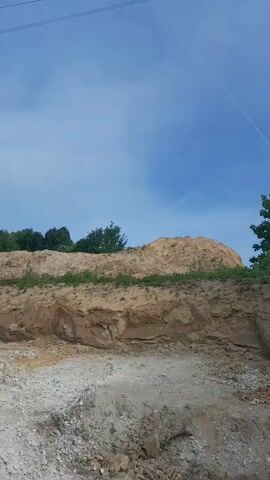 This media file depicts an outdoor scene, likely a construction site or a place where earth has been excavated. The foreground is filled with loose gravel and soil, indicating disturbed ground. Behind this, a large mound of sand or earth rises, with some green vegetation growing on its slopes and along its top. Further in the background, a line of trees forms a natural boundary against a bright blue sky.

The sky is clear with a few wispy white clouds, suggesting a sunny day, possibly late morning or early afternoon. There are no people or specific objects visible that would indicate a particular activity beyond the general impression of earthworks. The setting appears to be rural or semi-rural, given the natural landscape elements present. The overall mood is neutral, focusing on the landscape and the excavation. There is no visible text in the media file.