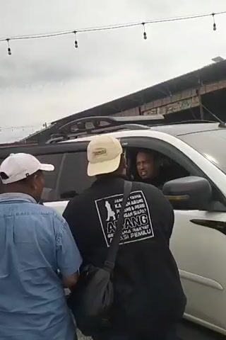 A group of three men are visible outdoors. One man is in the driver's seat of a white SUV, looking at the other two men. The man in the driver's seat is wearing a black shirt and a tan baseball cap. The other two men are standing outside the vehicle, facing the driver. One of these men is wearing a light blue collared shirt and a white baseball cap. The third man is wearing a black t-shirt with white text and graphics on the back, and a black cross-body bag. He is also wearing a tan baseball cap. The backdrop shows a cloudy sky and a building with a corrugated metal roof. String lights are visible overhead. The man in the black t-shirt appears to be speaking to the driver.