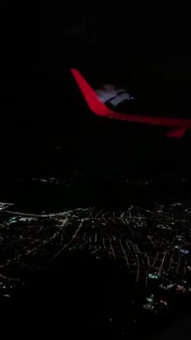 The video is a nighttime aerial view looking down at a city. The city lights are visible, illuminating the urban sprawl below. In the upper portion of the frame, there is an object that appears to be a plane or drone with blinking lights. The video continues to show the city from above, and then the wing of an airplane with a red tip becomes visible in the frame. The camera appears to be inside an airplane, looking out the window.