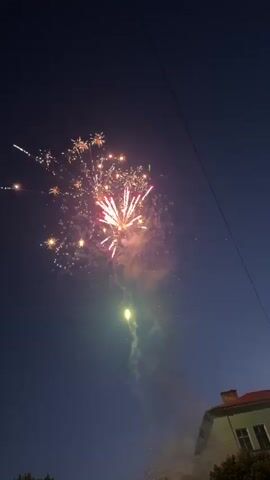 A vertical video shows several fireworks exploding against a dark blue evening sky. A single red firework trails upwards before bursting into a large, red and gold starburst, followed by multiple golden fireworks that erupt and twinkle, casting light on the surrounding smoke. A thin black power line runs diagonally across the upper right portion of the frame, and the top of a building with a reddish-brown roof and light-colored walls is visible in the bottom right corner.