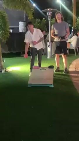 Two men are playing cornhole on an astroturf lawn outdoors at night. One man throws a pink cornhole bag at the board, which lands near the center. Behind the men, there is a black box truck, a few chairs and tables with people sitting, a few trees, and a tall patio heater. The men appear to be dressed casually.