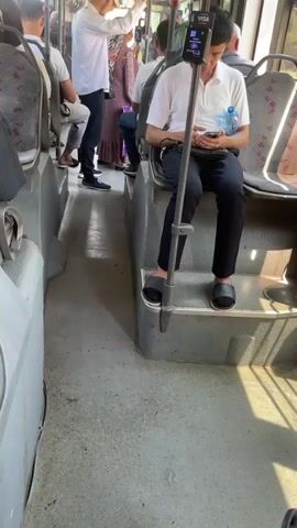 The video shows the interior of a bus with passengers. The camera is positioned looking down the aisle from the back of the bus towards the front. The floor of the bus is dirty. In the foreground, a man wearing a white short-sleeved shirt, dark pants, and black slippers is sitting on a raised step. He is holding a smartphone in his hands and looking down at it. A bottle of water is next to him. A metal pole with a device attached to the top, possibly a ticket scanner or information display, is in front of him. To his left, there is a row of seats. The aisle continues forward, where other passengers are visible. A man in a white long-sleeved shirt and a beige cap is standing near the front of the bus, holding onto a pole. He appears to be looking at something further forward. Further down the aisle, a woman in a patterned dress is standing. The windows of the bus show sunlight and trees or buildings outside. The seats on the bus have patterned upholstery. The lighting suggests it is daytime.