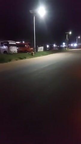 The video shows a dark street at night, illuminated by street lights. In the distance, there are parked vehicles and some activity visible. The camera is focused on a dark SUV, which is parked on the side of the road. A person is standing near the vehicle. The SUV's parking lights are on, adding a bit of illumination to the immediate surroundings. The overall atmosphere is dim and quiet.
