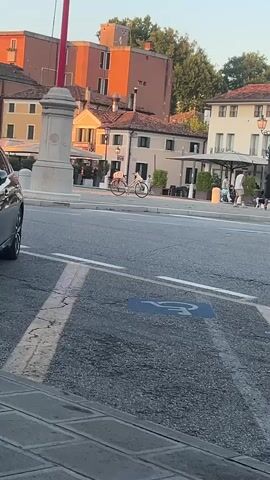 The video shows a street scene with buildings and people in the background. In the foreground, a disabled parking space is visible on the asphalt. A white bicycle is parked in front of a building. Several people are walking in the background near outdoor seating areas. A car is partially visible on the left side of the frame. The sun appears to be setting, casting an orange glow on the buildings.
