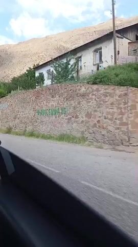 A first-person view from inside a vehicle shows a rough stone wall along a paved road. The wall has some graffiti on it, including what appears to be "EXPRESSLAB.KG" in white letters. Beyond the wall, there is a building with a white exterior and green shutters on the windows, situated on a hillside with sparse vegetation. The sky above is cloudy. The car is moving, indicated by the blur of trees on the right side of the frame and the slight motion blur of the scenery.