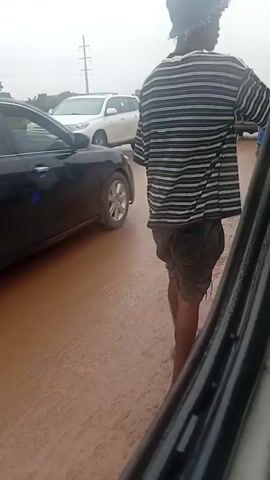 The video shows a road scene with several vehicles. In the foreground, a black car is partially visible on the right side. A person wearing a striped shirt and a hat is standing on the road on the right, facing away from the camera. A black sedan is driving on the muddy road. Further down the road, a motorcycle with three people on it is traveling. A red and yellow minivan is also on the road, and a white SUV is visible. The road surface appears to be muddy or wet. Power lines and some vegetation are visible in the background. The sky is overcast.