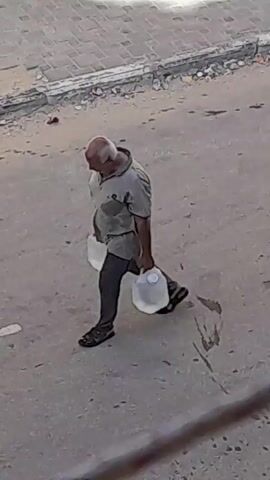 A bald, middle-aged man with gray hair on the sides of his head is walking from left to right across a paved area. He is wearing a short-sleeved collared shirt, dark pants, and sandals. He is carrying a large, clear plastic jug of water in his right hand. The jug appears to be full. The ground is rough and appears to be a road or sidewalk. There is debris in the background, including what looks like trash. The lighting suggests it is daytime. The video is grainy and appears to be taken from a distance.
