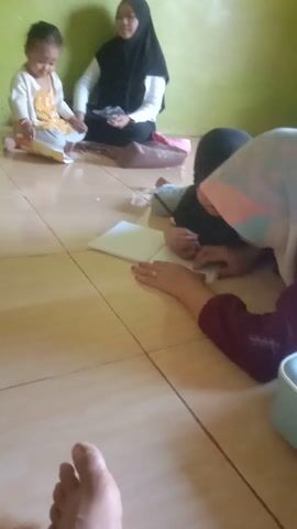 The video shows an overhead, tilted view of a room. In the foreground, a person's feet are visible. To the left, a woman wearing a black hijab and white shirt sits on the floor. Next to her, a toddler in a yellow and white outfit sits on the floor with papers. In the center, a person wearing a hijab with pink and blue accents and a maroon garment kneels on the floor, attending to a small child lying down. There are papers on the floor, a pink bag, and a light blue bag with a bow. The room has green walls and tiled flooring. Further back, more people are visible lying down or sitting, and a green bottle and some plastic-wrapped items are on the floor. The lighting is bright, possibly from a window or sunlight.
