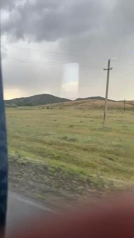 The video captures a scenic view of a rural landscape in Qarqaraly, Kazakhstan. The scene appears to be during the late afternoon or early evening, with dramatic, cloudy skies suggesting approaching rain. The terrain is characterized by rolling hills covered in dry grass and sparse vegetation.

Several objects are visible in the frame. A prominent wooden utility pole with wires stretches across the middle ground. In the distance, a cluster of white, dome-shaped structures, possibly yurts or modern glamping tents, dots the landscape. These structures indicate some form of habitation or tourist accommodation in this remote area. The foreground shows a glimpse of a road or pathway, suggesting the footage is captured from a moving vehicle.

The overall mood is one of vastness and natural beauty, tinged with the somberness of the overcast weather. The reflection on the glass (presumably a window) creates a faint, rectangular overlay on the scene, adding a layer of visual interest. No people are directly visible in the frame. The absence of immediate human activity emphasizes the tranquility and isolation of the setting. The landscape itself, with its open fields and distant hills under a brooding sky, evokes a sense of wilderness and a connection to nature.