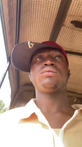 The video appears to be shot from the perspective of a person riding in what looks like a motorized tricycle, possibly an auto-rickshaw. The person is a young Black man wearing a two-toned baseball cap and a light-colored, collared shirt. He looks directly at the camera for the majority of the video. The background shows glimpses of a building, a utility pole, and other passengers. The camera moves slightly with the motion of the vehicle, creating a somewhat shaky view.