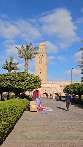 The video shows a sunny outdoor scene featuring a tall, ornately decorated minaret, which is a tower near a mosque from which Muslims are called to prayer. The sky is blue with scattered white clouds. In the foreground, there is a brick pathway, with various items including clothes, cardboard boxes, and other merchandise spread out, presumably for sale. A man in a dark jacket and pants is walking away from the camera along the pathway. Lush green bushes and palm trees are also visible. The call to prayer, or adhan, can be heard.