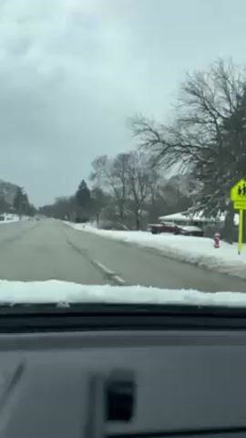 The video shows a view from inside a car driving down a snow-covered street. The sky is overcast. There are houses and trees on both sides of the road, also covered in snow. Other cars are visible on the road.