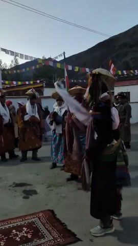 A group of people in traditional attire are gathered outdoors, possibly for a cultural event or celebration. Several individuals are wearing elaborate hats adorned with colorful ribbons and fabrics. Some are dressed in robes of varying colors and patterns, while others wear white scarves around their necks. In the foreground, a person in a dark robe and a wide-brimmed hat faces away from the camera, seemingly participating in a ritualistic dance. A patterned rug is laid out on the ground in front of them. In the background, buildings with colorful prayer flags strung across them are visible, with a mountain slope rising behind. The sky appears to be either dawn or dusk, with soft lighting.
