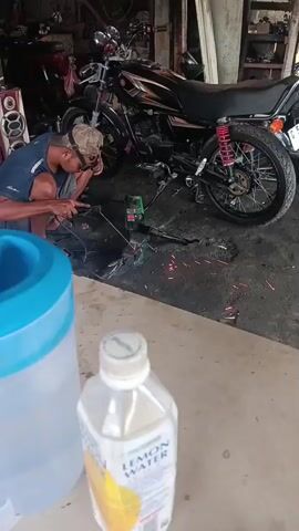 A man is crouched down, wearing glasses and a cap, and is welding a motorcycle frame. Sparks are flying from the welding tool. The motorcycle is dark colored with red accents on the rear suspension. A blue and clear plastic container is in the foreground to the left. A bottle of "Lemon Water" is in the foreground to the right. The scene appears to be a workshop or garage.