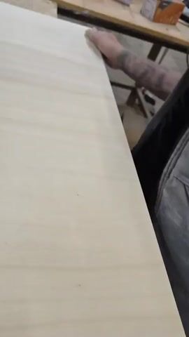 A person's hand is sanding the edge of a wooden board. The person is wearing dark clothing and has tattoos on their arm. The board is light-colored and appears to be unfinished. The scene appears to be a workshop or a construction site.