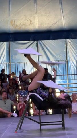 A young woman with long brown hair is performing a balancing act on a stage. She is wearing a sparkly black leotard and black ballet flats. She is reclined on a black padded surface that is on top of a black stool with red accents on the legs. Her body is inverted, with her head down and her legs extended upwards. She is balancing two large, flat, white objects with sparkly edges on the soles of her feet. The objects are triangular in shape. Her arms are outstretched, with her right arm bent at the elbow and her hand near her chest, and her left arm extended upwards. There are people in the background sitting in bleacher-style seating, watching the performance. The stage appears to be set up under a large tent or canopy. The lighting suggests it is daytime.