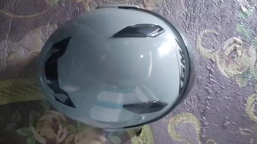 A light blue and black Kask helmet is positioned face-down, viewed from a top-down perspective, resting on a multi-colored patterned surface. The helmet has a glossy, reflective finish, showing faint blue and white reflections. It features several black ventilation openings on its top surface and a black rim along one side where the brand name "KASK" is visible in white letters. The underlying surface displays a dark green, brown, and light floral pattern.