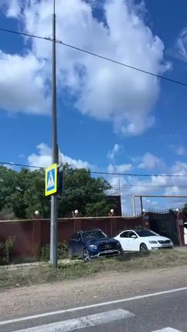 The video captures a view from a vehicle traveling along a road. The sky is blue with scattered white clouds. On the left side of the road, there is a house with a dark fence and two parked cars, a white Skoda and a white minivan. Further down the road, on the right side, there are more houses, some with distinctive roofs, fences, trees, and utility poles with power lines. The road appears to be paved. The video moves horizontally, showing a continuous panorama of the roadside scenery.