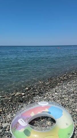 The video captures a sunny day at a pebble beach. The camera is positioned close to the ground, focusing on a colorful inflatable ring in the foreground. Beyond the ring, the clear blue water of the sea stretches to the horizon, with several people swimming and floating. On the beach, a young boy in a black t-shirt and shorts walks towards the water, carrying a bag. Sunbathers are visible relaxing on lounge chairs under a green umbrella in the background. The overall scene depicts a typical summer day at a seaside resort.
