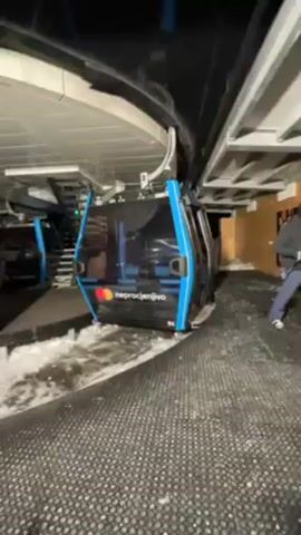 A ski gondola with black and blue trim sits in a station, which is a covered area with a black ceiling and a black rubber-like mat on the floor. The gondola has the word “NEPROCJENJIVO” printed on its side, along with a Mastercard logo. Patches of snow are visible on the floor around the gondola.  A person wearing a dark coat and a hat enters the gondola. A poster featuring food is visible on the wall to the right of the gondola.