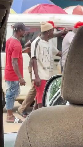 The video shows a group of people standing near a white van. A man in a red t-shirt and blue jeans is on the left, facing away from the camera. He is wearing a black baseball cap and sandals. In the center, a man in a cream-colored t-shirt with a graphic print and red shorts is looking to the right. He is wearing a light-colored beanie and jewelry. To his right, a person in a dark shirt and red cap is facing the camera and gesturing with their right hand. Behind them, more people and colorful umbrellas are visible. The view is from inside a vehicle, with the headrest and interior of the car in the foreground. The environment appears to be outdoors, possibly a street or market.