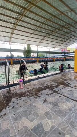 The video shows an indoor swimming pool with a tiled floor and a metal roof. Several people, including adults and children, are in the pool, some of whom are swimming or playing in the water. One person is at the edge of the pool, appearing to be observing. There are metal handrails near the edge of the pool. A pink water bottle is visible near the handrails. The water is choppy in some areas, suggesting activity. The environment appears to be a public or recreational swimming facility.