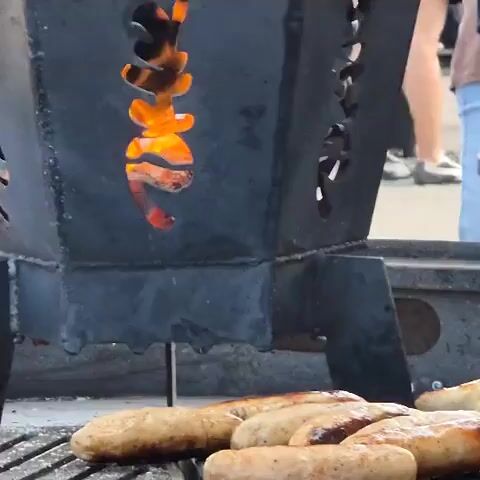 The video shows a close-up shot of sausages being grilled. The sausages are arranged on a metal grate over a fire, with flames visible through a decorative opening in the grill. In the background, people are standing around, some appearing to be watching the grilling. The environment looks like an outdoor gathering or event. The focus is on the cooking process, with the sausages appearing to be browned and slightly charred.