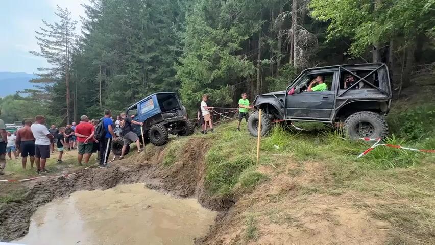 A group of people watch two off-road vehicles engaged in an off-road competition. The vehicle on the left is blue and appears to be stuck in mud at the bottom of a hill. The vehicle on the right is black and is on top of the hill, with a tow rope attached to the blue vehicle. Several people are gathered around the blue vehicle, some appearing to be assisting. The environment is wooded with a muddy track and a body of water visible.