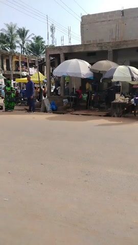The video shows an active street scene in Awka Etiti, Nigeria, during the daytime under clear, sunny skies. The setting is a bustling commercial area or market with light brown, unpaved ground. Numerous people are present, walking, standing, and congregating.

In the foreground, a man in a blue outfit and a woman in a green patterned dress are standing on the left side of the street. Another man walks from left to right past them. Further down the street, several market stalls are visible, characterized by large white and beige umbrellas providing shade for vendors and customers. Many people are gathered under these umbrellas, suggesting active commerce.

Buildings, some appearing to be concrete structures under construction with exposed frames and others established, line the street. Palm trees are visible in the background, and power lines crisscross overhead. A yellow Keke Napep (auto-rickshaw) drives by at one point. Later, a young woman with long braids, wearing a black short-sleeved top, a grey or olive green apron-style dress, and black leggings, walks past the camera from right to left. A black bag is placed on the ground near the man in blue, and a blue sign with indistinct text is also visible. The overall scene depicts the lively daily activity of a local community market.