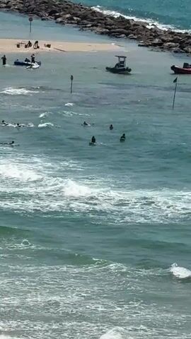 This video shows an aerial view of a beach and ocean scene. In the foreground, gentle waves are breaking and rolling towards the shore. Several people are visible on surfboards in the water, some paddling and others sitting on their boards. In the background, a breakwater made of large stones separates a calmer, shallower area of water from the open ocean. A sandy beach is visible behind the breakwater, with a few people and a jet ski on the sand. Several boats, including a small inflatable boat and a larger motorboat, are anchored or moored in the calmer water. Two black poles are sticking out of the water, possibly marking a boundary or hazard. The water is a turquoise blue, and the sky appears clear and bright, suggesting a sunny day.