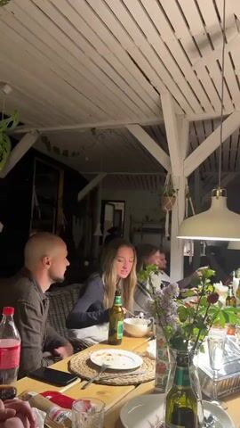 A group of people are sitting around a long wooden table under a pergola, eating and drinking. The pergola has a white ceiling and is decorated with hanging plants. The table is cluttered with plates of food, glasses, bottles of beer and soda, and a vase of purple flowers. Two large pendant lights hang from the ceiling. A man is sitting at the near end of the table to the left, a woman is next to him, eating spaghetti. Other people are visible further down the table, eating, drinking, and using their phones. The overall atmosphere appears casual and social.