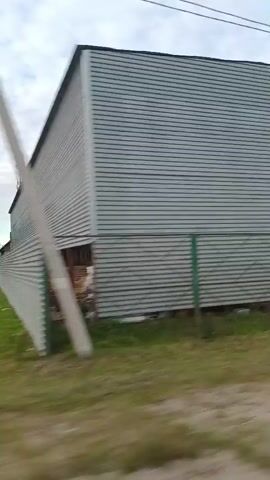 The video shows a view from a car driving down a road. The scenery is rural, with trees and tall grass. There are power lines visible in some shots, as well as a large industrial-looking building. The sky is cloudy. The car passes by a traffic light and signs.