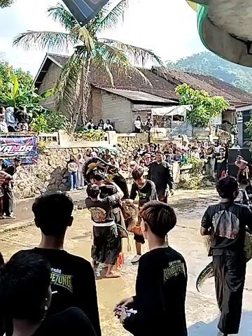 The video shows a traditional Indonesian martial arts performance in a village setting. Two performers in black outfits engage in a physical routine on a wet, concrete surface, observed by a large crowd. Some members of the crowd are wearing black shirts with similar designs, possibly identifying them as part of the performing group or organizers. 
Behind the crowd are various houses, a stone retaining wall, and a banner with the word "SIYANDA" on it. A palm tree is visible in the foreground, adding to the rural ambiance.
The performance takes place in a muddy area, indicating recent rain or the presence of a shallow body of water. One of the performers falls onto the ground, while the other continues to move dramatically. The overall scene indicates a community event showcasing traditional arts and culture.