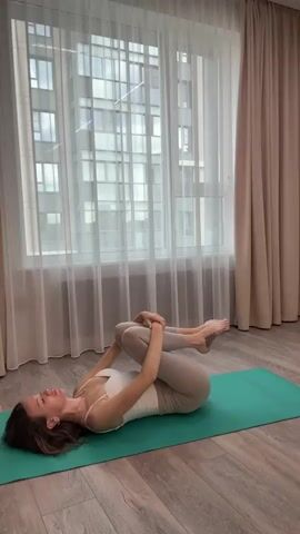 A woman is lying on her back on a green yoga mat in a room. She is wearing a white tank top and beige leggings. Her knees are bent and drawn up to her chest, and she is holding them with her hands. Her head is tilted back, and her eyes are closed. Her hair is dark brown and falls around her shoulders.

The room has light-colored wooden floors. Behind the woman, there is a large window with sheer white curtains and thicker beige curtains on either side. The window offers a view of a modern building with many windows. The lighting suggests it is daytime. The woman's posture indicates she is likely engaged in a stretching or relaxation exercise, possibly a yoga pose. There is no visible text in the media file. The setting appears to be an indoor space, possibly a home or a yoga studio, in Chashnikovo, Russia.