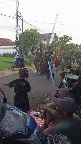 The video depicts a street scene where a small parade seems to be taking place. In the foreground, a young child wearing a black t-shirt stands next to a blue moped, looking towards the road. A man in a green t-shirt and a baseball cap sits nearby, with another individual whose head is partially obscured by a hijab. The moped’s speedometer is visible.

In the middle ground, two women in elaborate costumes and headdresses are walking down the street. One costume is predominantly gold, while the other appears to be red and green. They are accompanied by a man holding a long blue pole.

In the background, other individuals are present, suggesting a gathering or parade. A man on a moped is driving in the opposite direction. A white flag with text in an Asian script flutters. The setting is a rural or semi-rural area, with a rice field visible on one side and houses on the other. Telephone poles and wires cross the sky. The lighting suggests an overcast day.