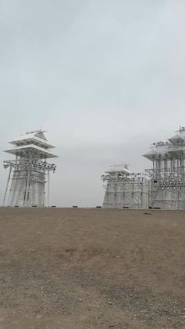 The video shows a large open space with three large metal structures resembling pagodas in the distance. The ground is covered in gravel and dirt. The camera pans down to show a person wearing blue jeans and brown cowboy boots walking on the ground. The sky is overcast and gray. There is faint audio of people speaking.