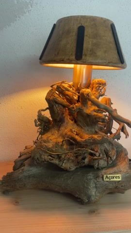 A still life shot showcases a lamp constructed from driftwood. The base of the lamp is a gnarly tangle of weathered wood, surmounted by an elaborate, root-like structure. A copper pipe extends upward from the wooden mass, supporting a lampshade of rough-hewn material with evenly spaced black strips. A small, rectangular plaque bearing the inscription "Açores" is affixed to the lower section of the base. The lamp is illuminated, casting a warm glow on the wooden components and creating a diffused light pattern on the stark white wall behind it. The lamp is positioned on a light-colored wooden surface. There is no one in the frame.