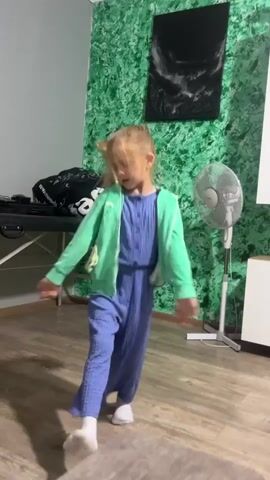 A young girl with blonde hair tied in a ponytail, wearing a blue jumpsuit and a green zip-up hoodie, dances energetically in a room. The room has a green textured wall behind her with a black framed picture and a white cabinet. A white fan is visible to her right. To her left, there is a black table and a large bag. The floor is wooden. The girl moves her arms and spins, her movements appearing joyful.