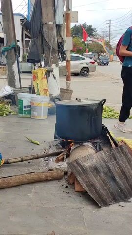 A large black pot sits atop a makeshift wood-burning stove on a paved sidewalk beside a road. The stove is constructed from wood, bricks, and a piece of corrugated metal. Smoke rises from the fire beneath the pot. Several ears of corn are scattered around the pot and near a wooden pole propped against a utility pole.  Two buckets, one green and one yellow, sit nearby, along with a large sack and other sundry items. In the background, cars and motorcycles travel along the road. Flags and pennants decorate the street. The scene appears to be an outdoor cooking setup alongside a busy street in a developing area.