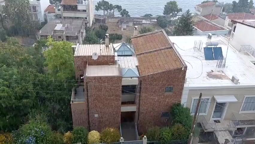 An aerial view shows a coastal town under heavy rainfall. The camera pans over a multi-story brick building with a brown tiled roof and a glass skylight. Adjacent to it, a white-roofed building features solar panels. Beyond these structures, houses with red tiled roofs are visible, nestled amongst lush green trees. In the background, the sea stretches out under a cloudy sky, with a large cruise ship docked in the distance. The heavy rain is evident throughout the video, creating a sense of a stormy atmosphere.