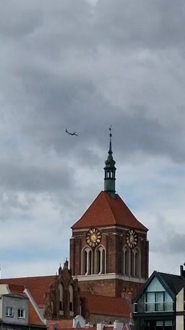 An airplane flies past a church steeple under a cloudy sky. The church steeple is tall and made of brick with a red roof. It has two clock faces visible. Below the steeple, there are parts of other buildings, including a modern building with balconies and large windows. The airplane is small in the frame and appears to be in flight. The sky is filled with gray clouds.