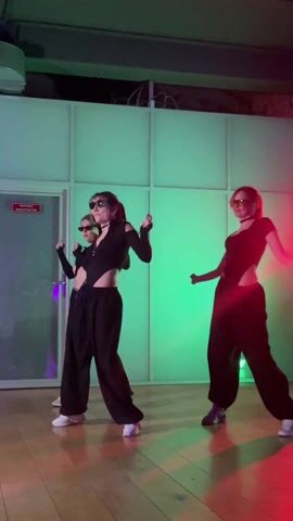 The video shows three young women dancing in a studio with white paneled walls. They are all dressed in black clothing: fitted tops and loose-fitting pants, and are also wearing sunglasses. The lighting alternates between green and red. The women move in unison, performing synchronized dance moves. The floor is wooden. A door with a red sign is visible on the left side of the frame. A ventilation unit can be seen near the top of the frame.
