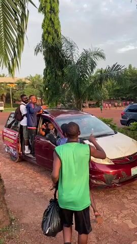 Several people surround a maroon minivan parked on a brick surface surrounded by trees. The front passenger door is open, and two men are standing on the vehicle’s floor. One man wears a black baseball cap, a blue and black striped shirt, and a red purse. The other is wearing a blue jacket and pointing to his right. A third man in a green tank top stands facing away from the camera with his right arm raised, carrying a black trash bag. Another man stands near the open car door wearing a white collared shirt, khaki pants, and a backpack. A driver can be seen sitting in the driver’s seat. Some bystanders are visible in the background near a building. The words "EXTRAMATIC ENTERTAINMENT" are on the side of the car.