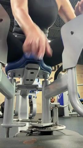 A woman with blonde hair wearing a black tank top and black leggings is sitting on a weight machine in a gym, performing a leg abduction exercise. The machine is silver and blue, and the woman is pushing the leg pads outwards against resistance. The camera angle is low, looking up at the woman and the machine. The gym environment is visible in the background, with other machines and workout equipment.